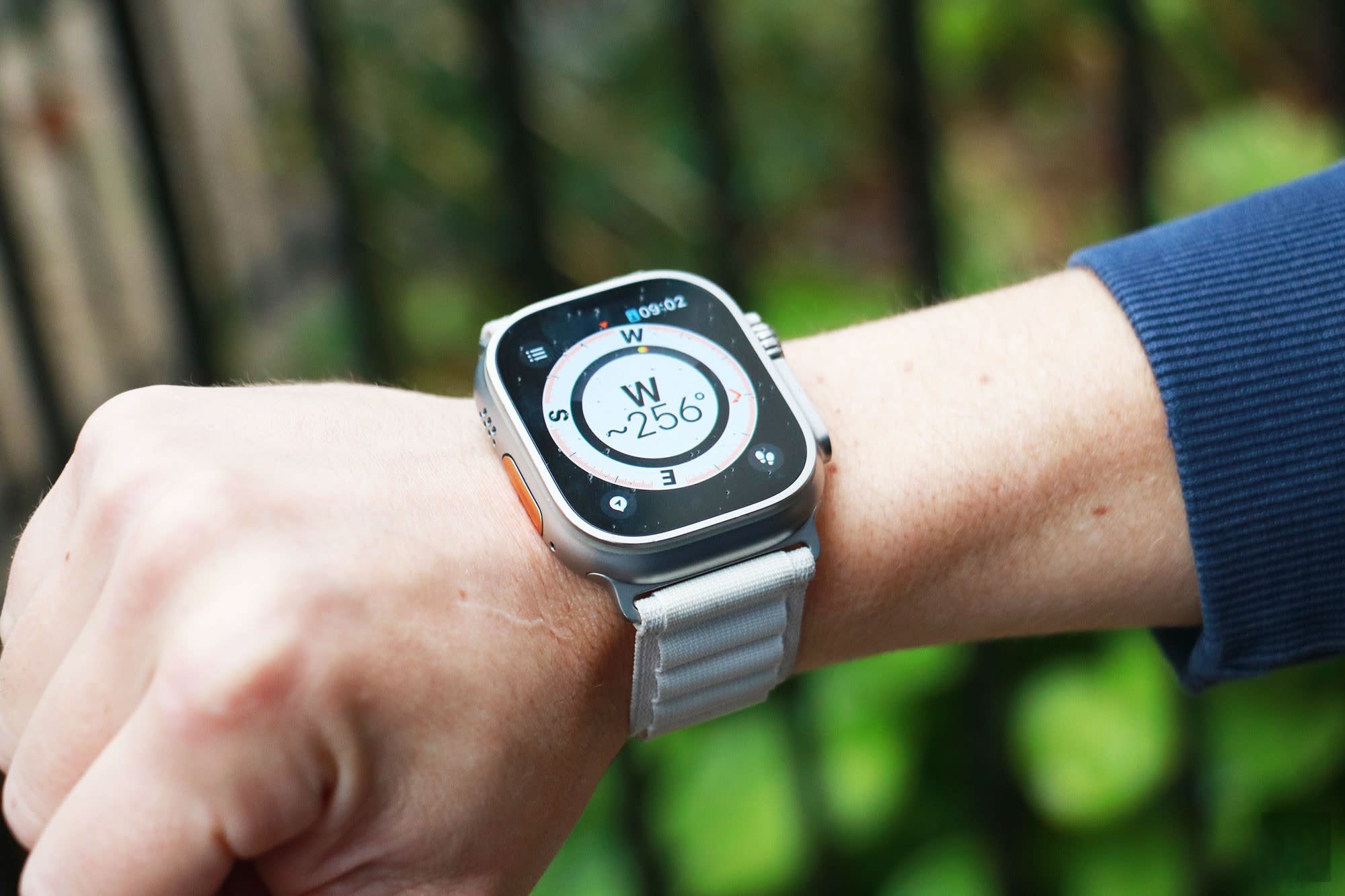 From Phonegram: A smartwatch displaying a compass worn on a person's wrist, pointing west at a 256-degree angle. Amidst slightly blurred green spaces and dark bands, this scene echoes recent discussions about the Apple Watch.