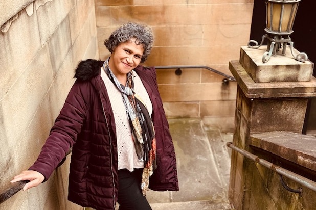 A women with short curly grey hair wearing a pruple jacket smiles while standing on a staircase. 