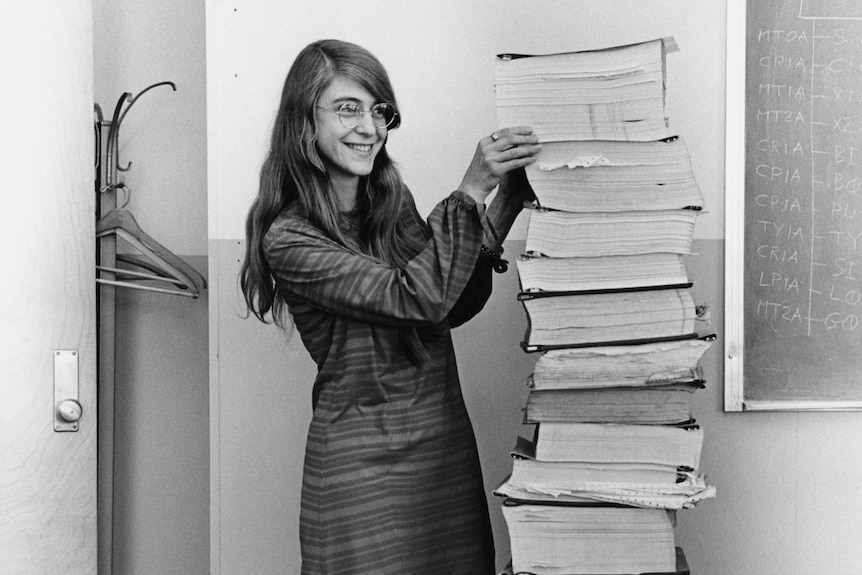 Black and white photo of woman with long hair, glasses and a long-sleeved dress next to a stack of bound papers as tall as her.