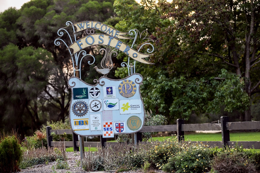 A metal roadside sign reads 'Welcome to Foster' with a shield below displaying logos of community organisations.