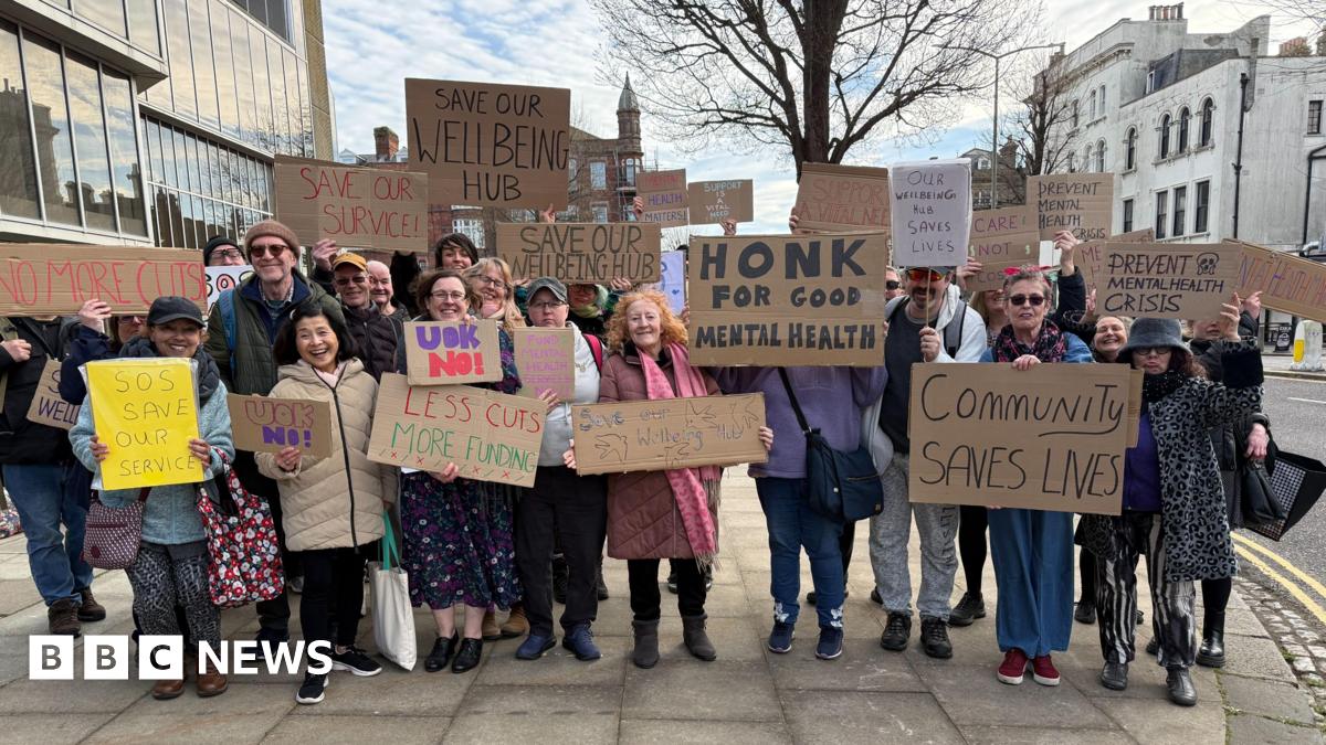 A group of people stand outside a building on a street. They are holding home-made placards in support of a mental health service which risks being cut.