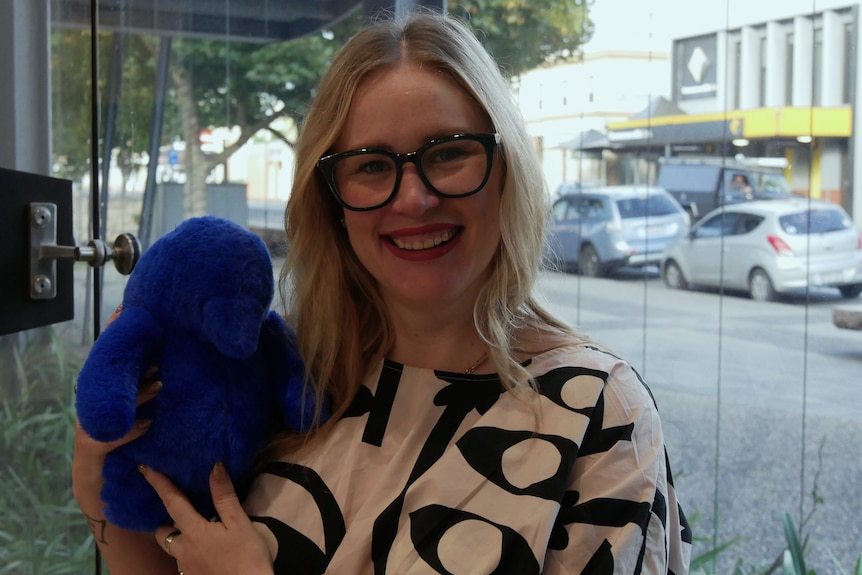 A woman wearing a black and white top and glasses holding a plushy blue toy.