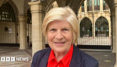 Sally Keeble is wearing a dark suit and red shirt. She has short blonde hair.