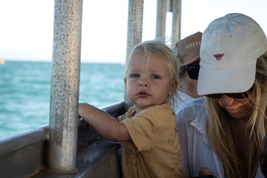 A blond baby looks into camera, arms up on the side-railing of a boat.