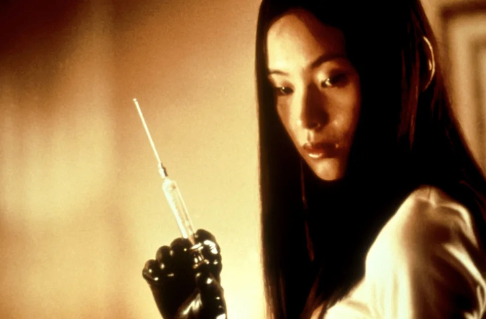 A woman holding a syringe with a focused expression, wearing black gloves in a dramatic setting