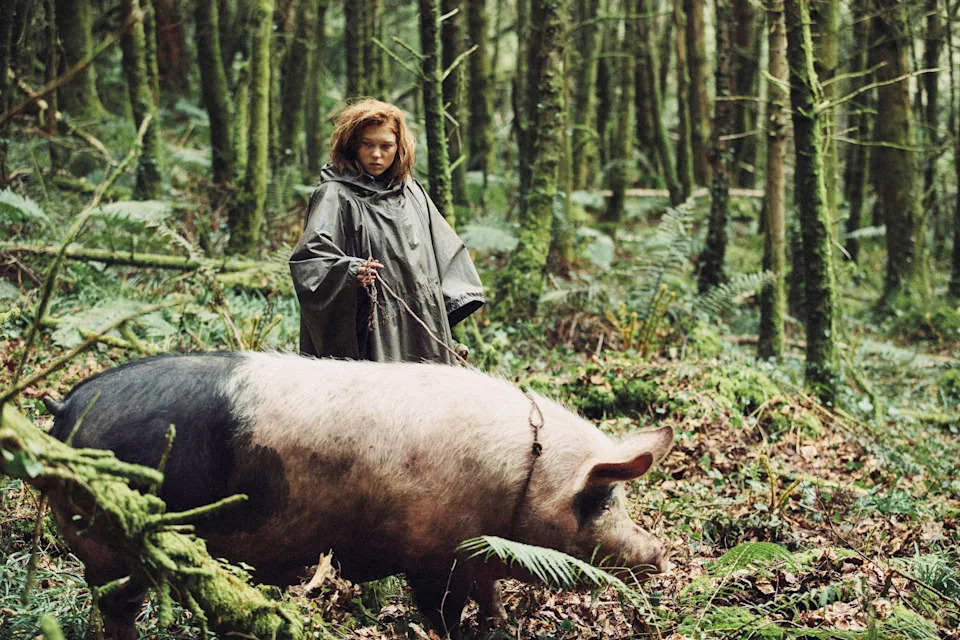 Person in a forest leads a large pig. The person is wearing a cloak and appears focused. The scene is dense with trees and foliage