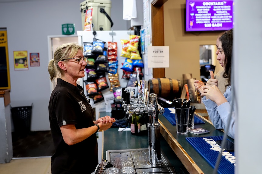 Woman wearing glasses and black shirt stands behind bar talking to two young customers 