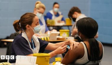 A nurse administers a vaccine does to a girl who is seated and faces away from the camera. The rest of the image is blurred but others can be seen also receiving their dose