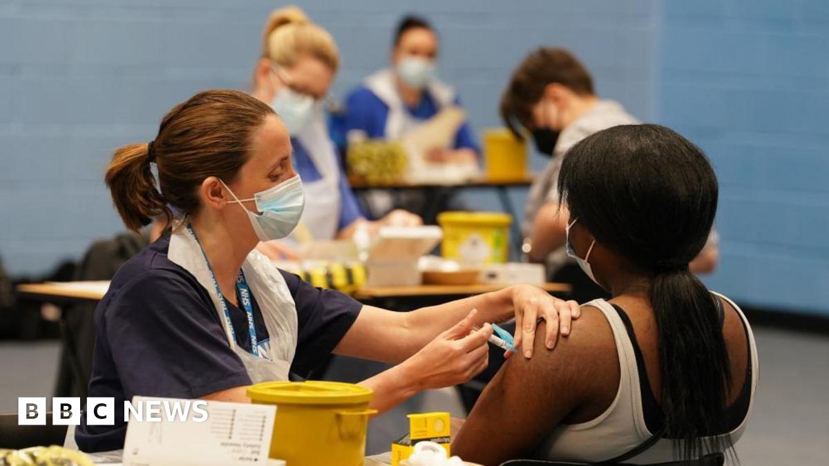 A nurse administers a vaccine does to a girl who is seated and faces away from the camera. The rest of the image is blurred but others can be seen also receiving their dose