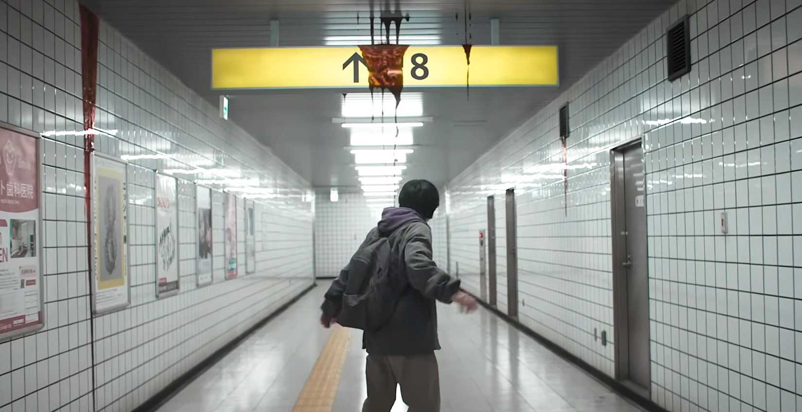 A young man in a brightly lit subway corridor begins to back away as blood drips from the yellow Exit 8 sign overhead in Exit 8