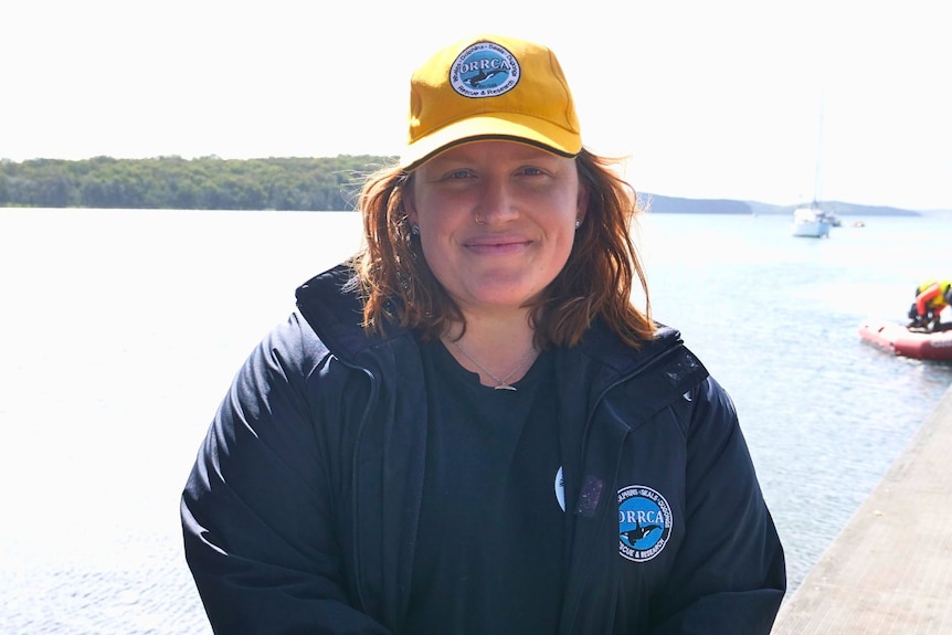 A woman wearing a yellow cap and dark blue jacket. A body of water is in the background.