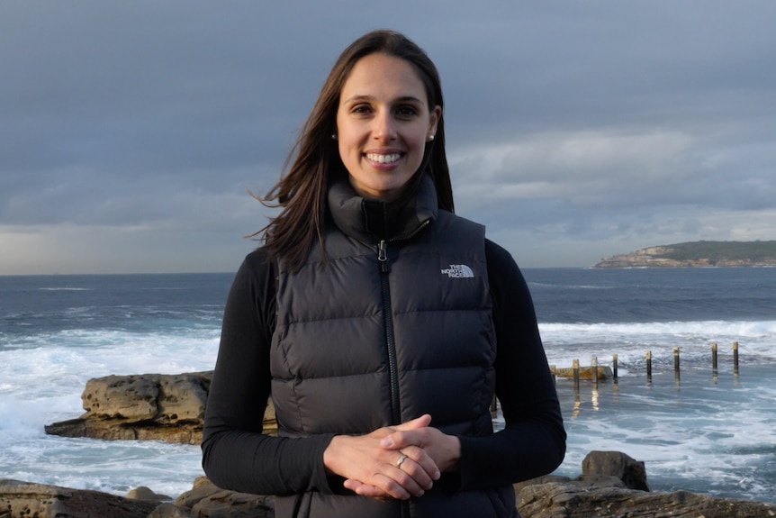 Vanessa Pirotta wears a black top and black puffer vest and stands in front of rocks and sea water.  