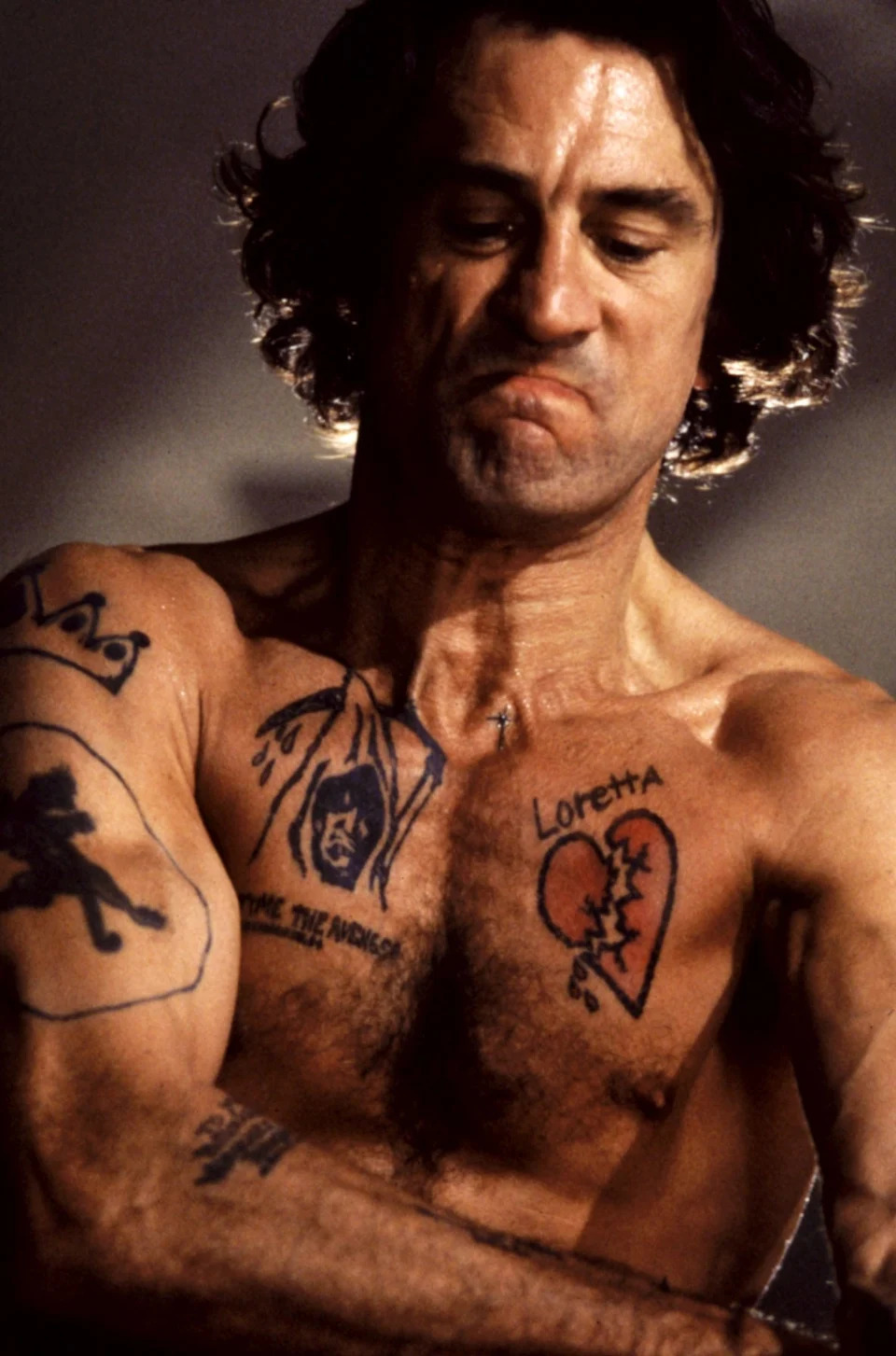 A shirtless person with various tattoos, including a broken heart and a hooded figure, is looking down with a serious expression