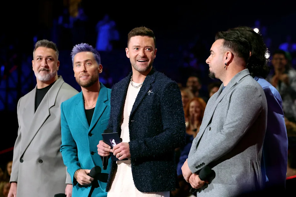 A group of four people in stylish suits smiling at an event stage, holding microphones, with a lively audience in the background