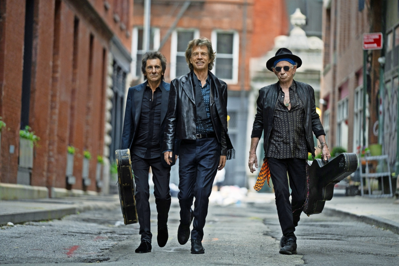 The Rolling Stones, Mick Jagger, Ronnie Wood, and Keith Richards, walk down an urban street carrying guitar cases.
