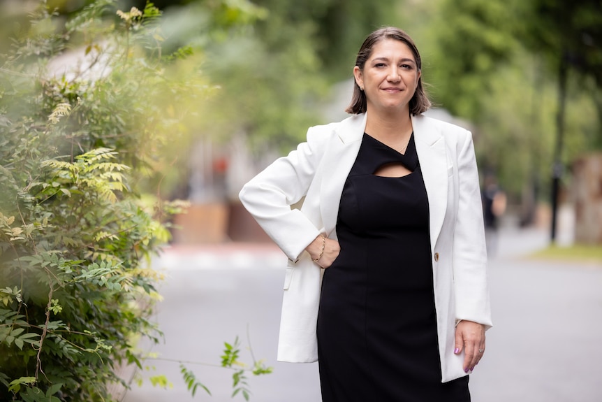 A woman with brown hair wearing a black dress and white blazer stands with her hand on her hip