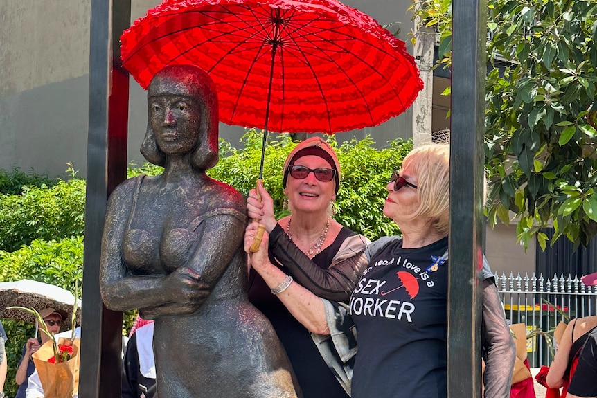 Two older women stare at a bronze statue of a woman leaning against a door. 