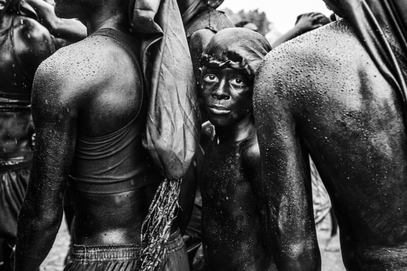 A group of people covered in dark body paint stand closely together; at the center, a child looks directly at the camera amid the crowd, droplets of water visible on their skin and clothing.