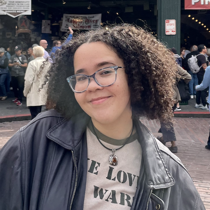 A person with curly hair, glasses, and a leather jacket smiles outdoors in a busy market area with people in the background.
