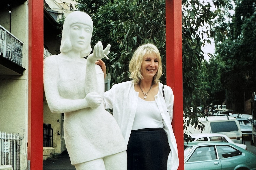 A woman stands next to a white scuplture of a woman smoking. 