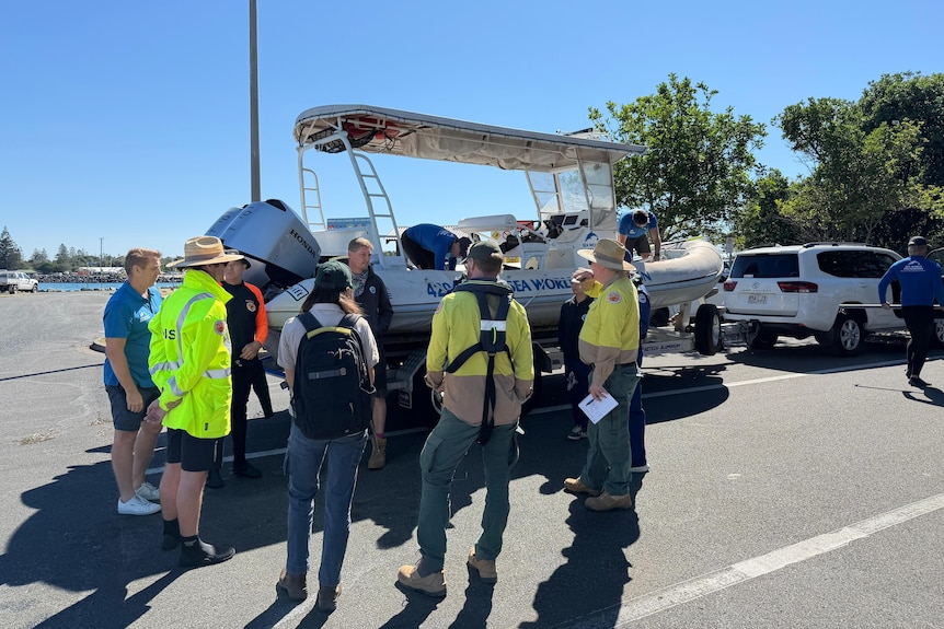 Group of people in uniforms stand next to Sea World boat on a boat trailer in a car park.