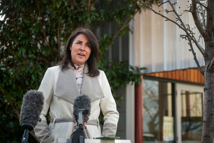 A woman with short dark hair wearing a white coat speaks into microphones outside a silver building and green trees.