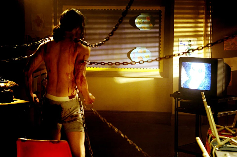 Person in a dark room connected to chains, facing a static-filled TV. Sparse lighting from a window