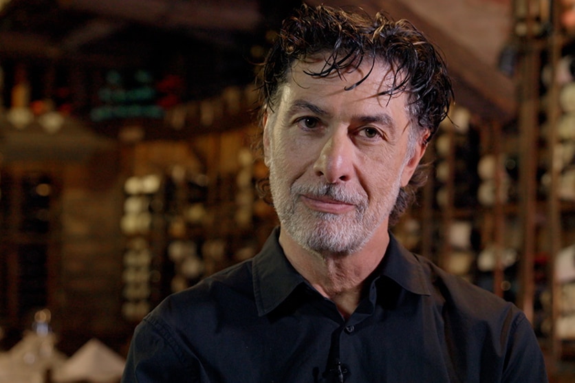 A man with a grey beard and black shirt stands in front of a wine cellar. The wine bottles are out of focus. 