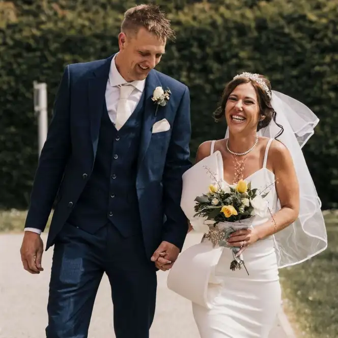 The bride and groom smile while walking outdoors, holding hands, with the bride holding a bouquet of yellow and white flowers.