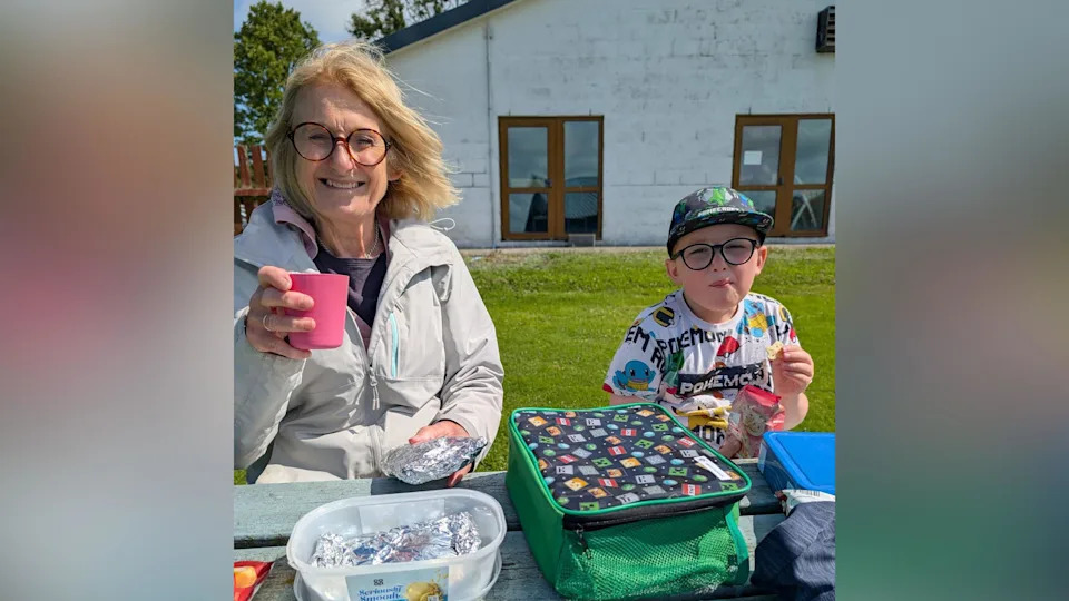 Helen sat with her grandson on a picnic bench in the sun. They are both eating a packed lunch on the table in front of them and smiling. Helen holds a pink plastic cup in the air, she is wearing a grey raincoat. 