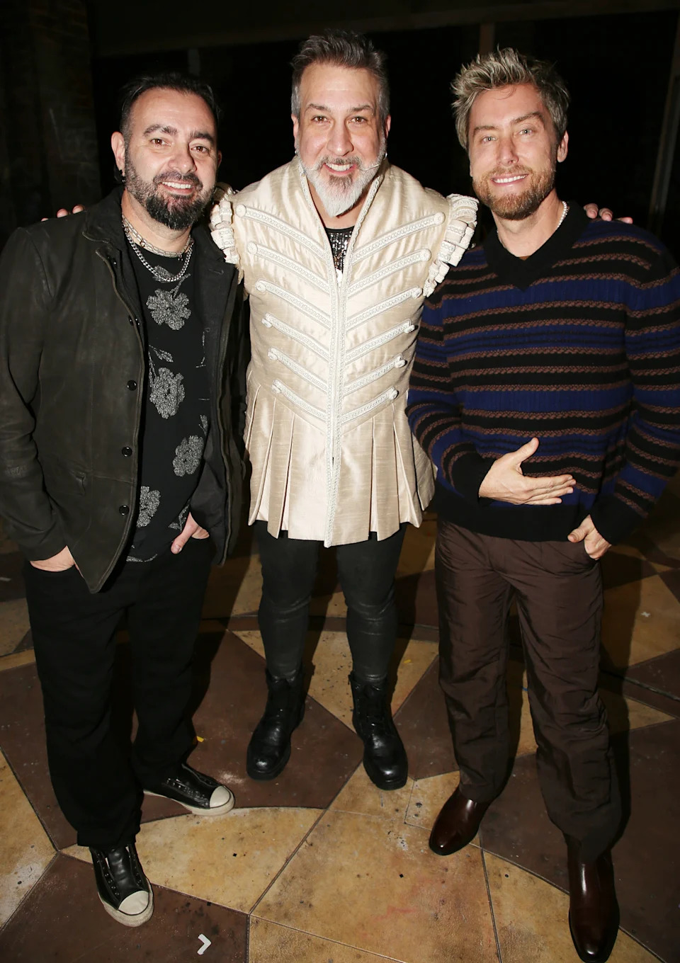 Three men pose together, with the central figure wearing a unique jacket with pleats and patterns, flanked by casually dressed companions