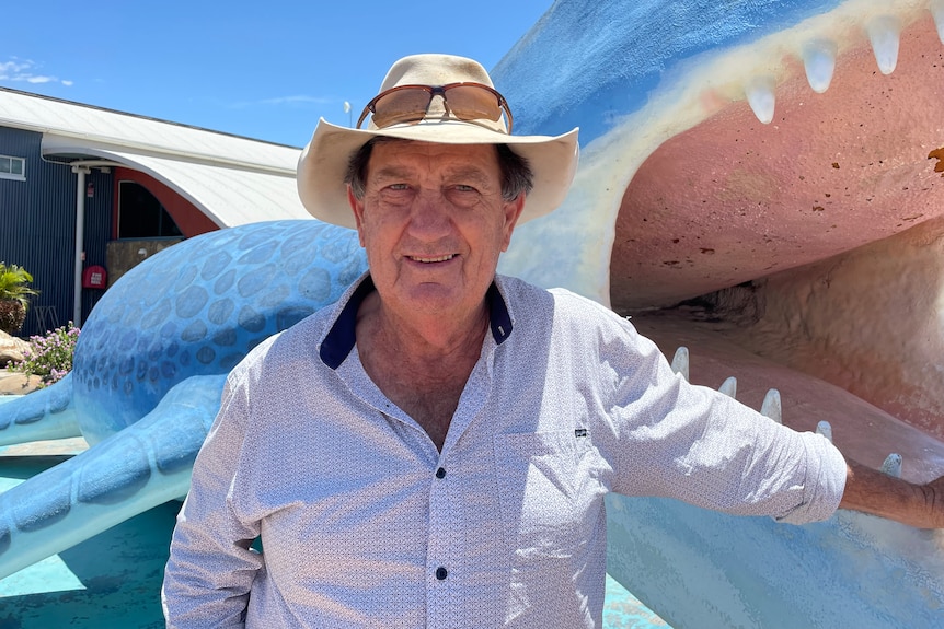 Man in shirt with hat on stands in front of a blue marine dinosaur sculpture.
