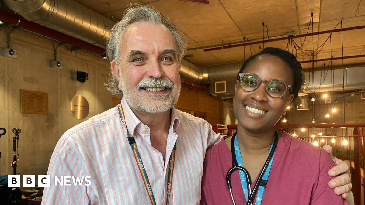 Ashley Brown is seen in a pink and white striped shirt with a rainbow NHS lanyard stands next to Mainga Bhima in burgundy medical scrubs with a stethoscope around her neck. They are both smiling in an indoor setting with industrial-style lighting in the background.
