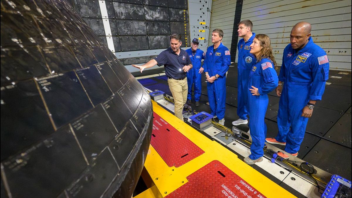 six people, including four nasa astronauts wearing blue flight suits, inspect the black bottom region of a conical space capsule inside a ship's well deck at sea