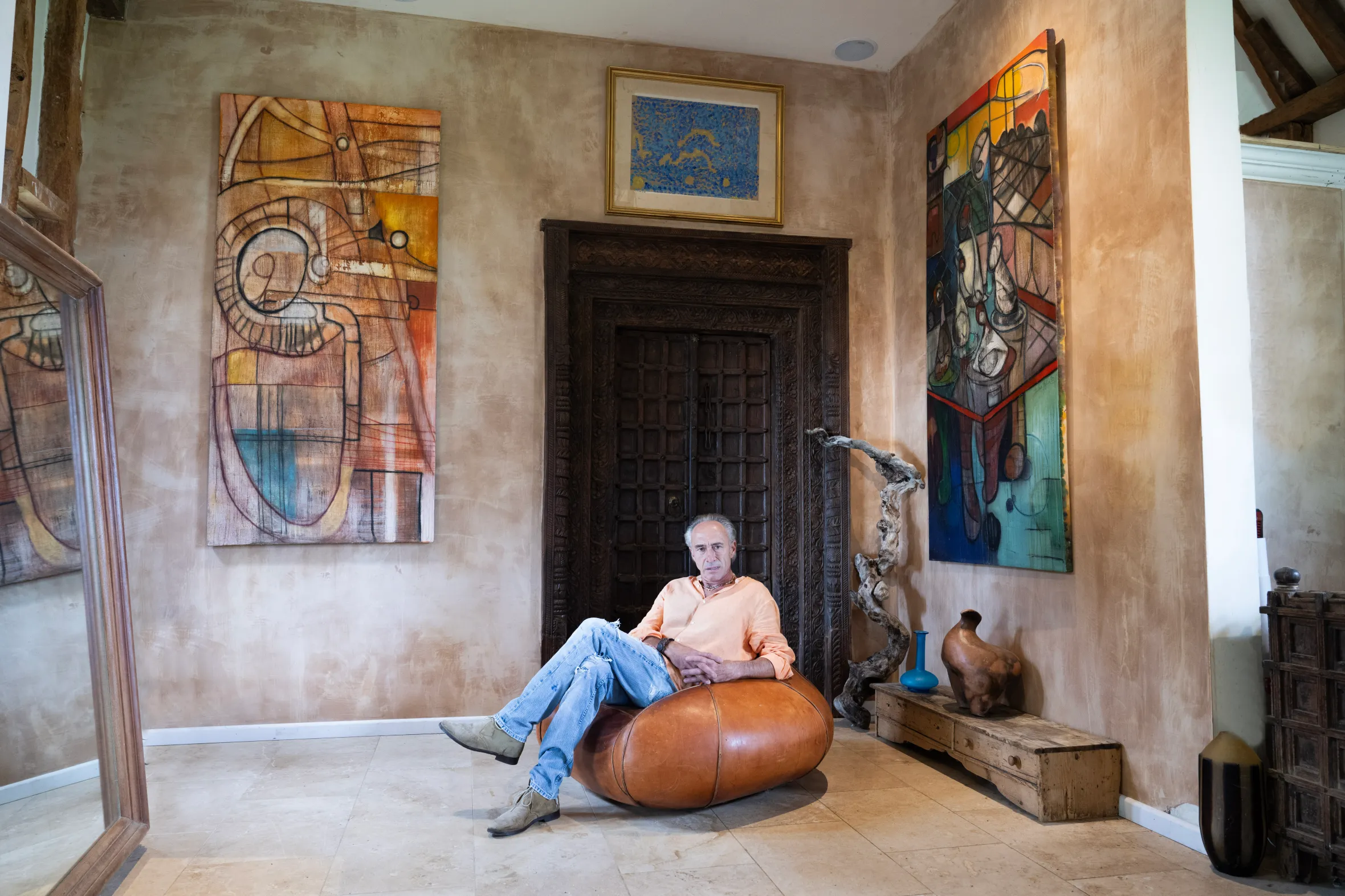 Clinton Pugh sitting on a leather pouf, framed by Sacha Jafri's "Mechanics of the Soul" and "Reflections of the Soul" paintings.