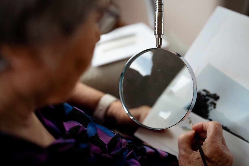 Woman sits on chair, using a magnifying glass to paint.