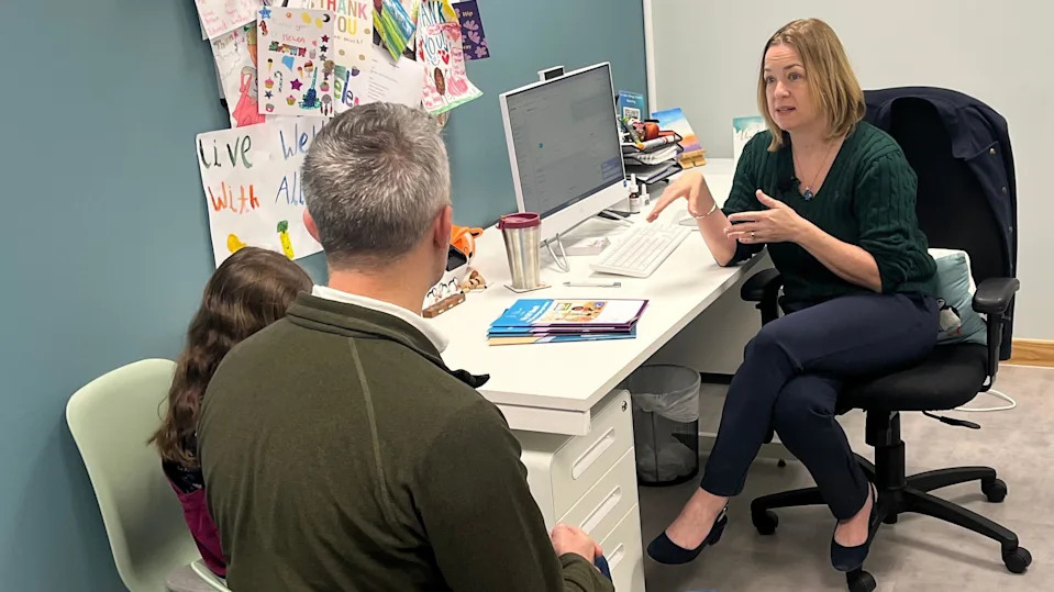 Dr Helen Evans Howells, sitting in office chair, talking to patients. Scott, a father wearing a green jumper, sits next to his young daughter Carey with long brow hair in consulting chairs, with their backs to the camera.