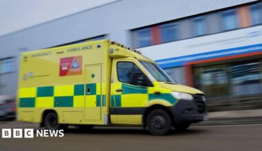 A photo of a green and yellow ambulance driving along a road, there is a blurred hospital in the background