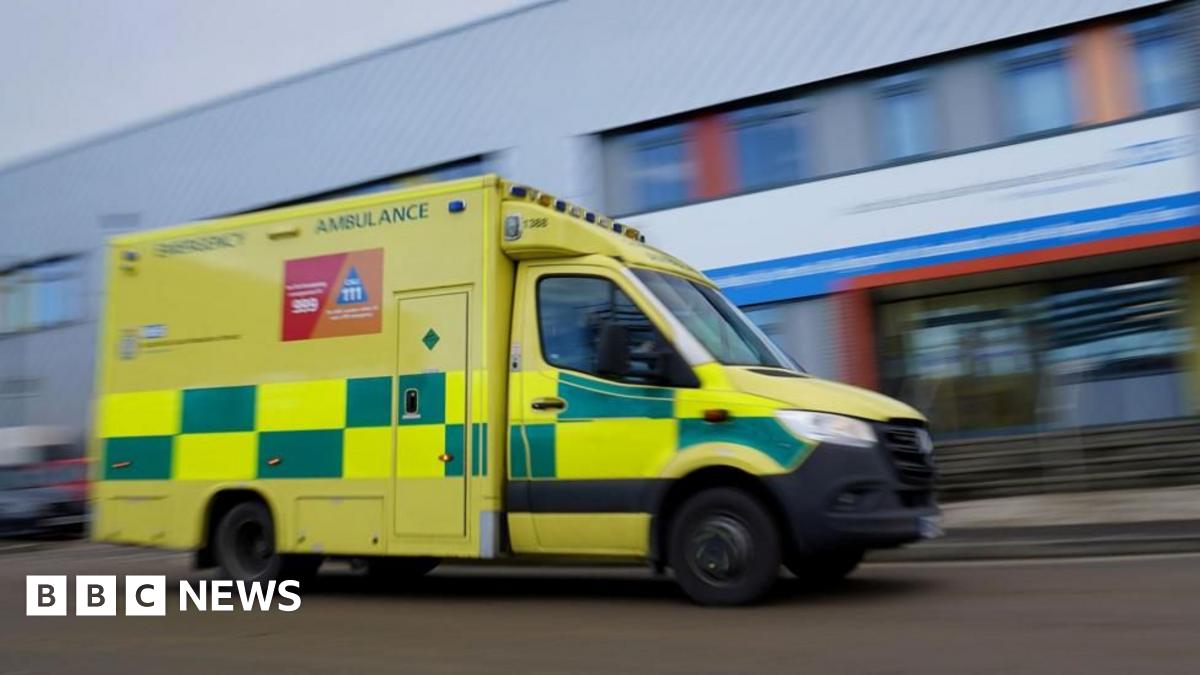 A photo of a green and yellow ambulance driving along a road, there is a blurred hospital in the background
