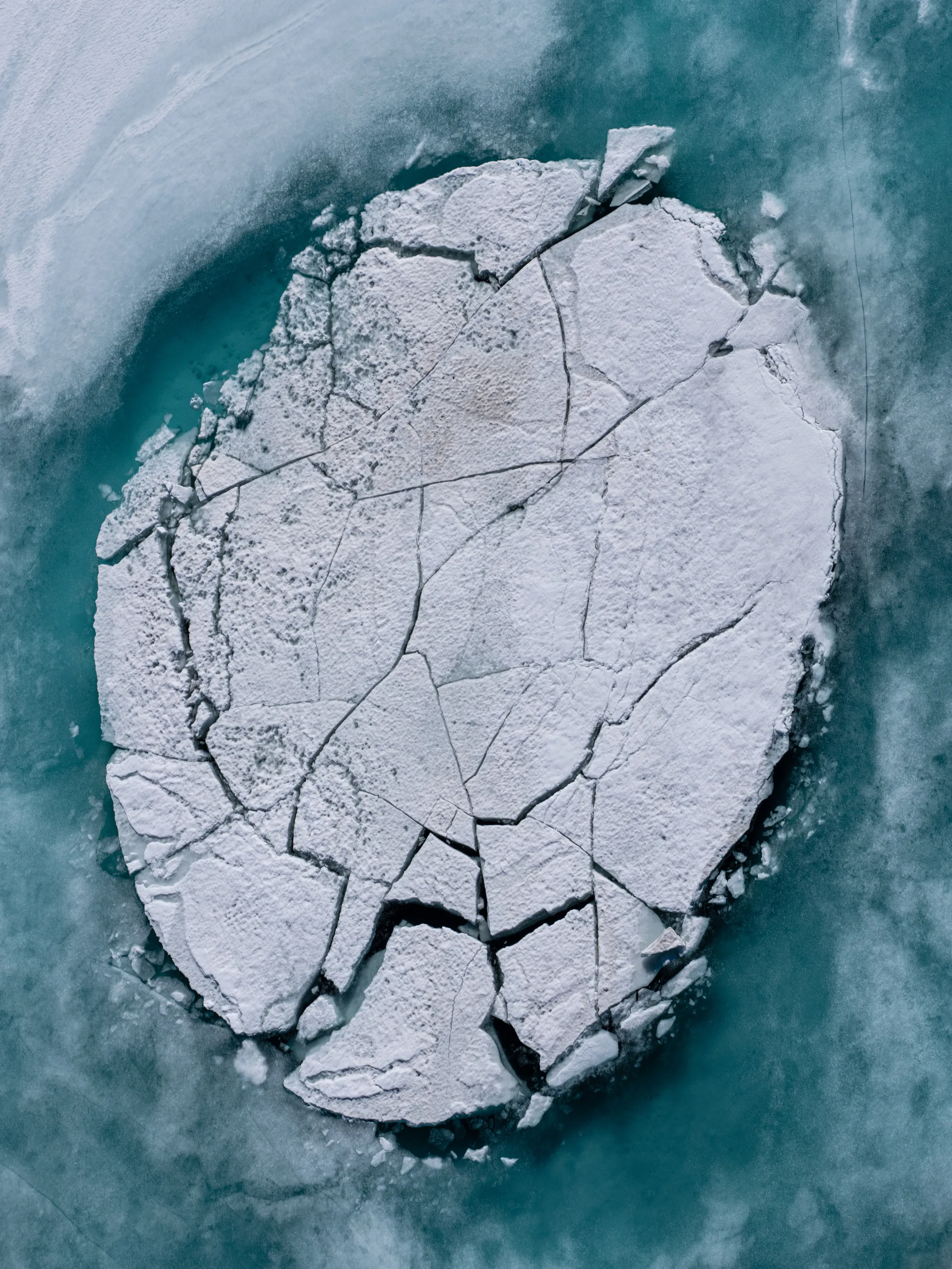 Aerial view of a large, circular ice floe fractured into many pieces, floating on a turquoise lake in Iceland.