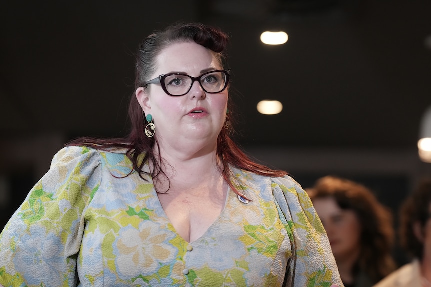 A person with dark hair wears a floral dress while speaking in a room.