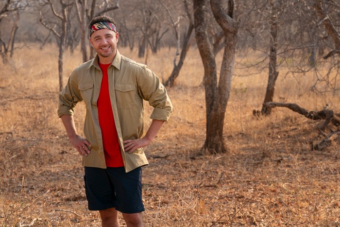 I'm-a-Celeb-South-Africa-2026 Adam Thomas stands with his hands on his hips smiling ahead in a safari landscape.
