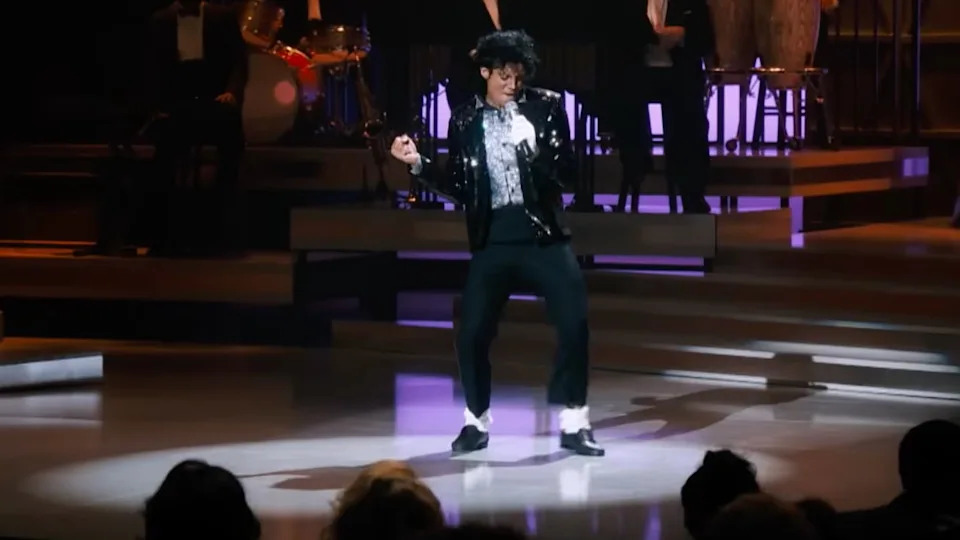 A performer on stage in a Michael Jackson-inspired outfit, including a sequined jacket, white socks, and black shoes, dancing energetically.