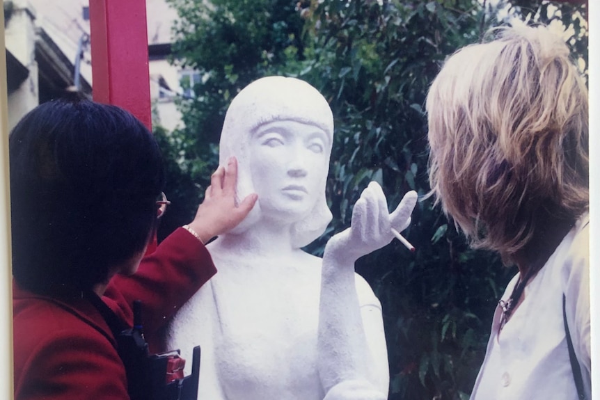 Two women touch a white cement statue of a woman smoking.