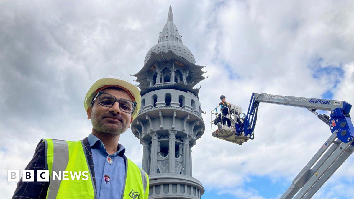 A man wearing a white safety helmet and tabard with the top of an ornate grey tower in the background.