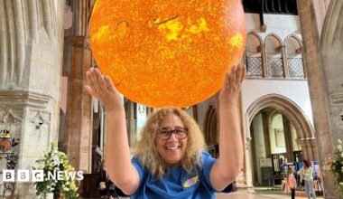 A woman with shoulder-length blonde hair is smiling and has her arms up in the air, creating the illusion that she is holding the large model of the Sun which is above and behind her. She is wearing tortoiseshell glasses and a blue top with an eye logo on it. She is standing inside a church.