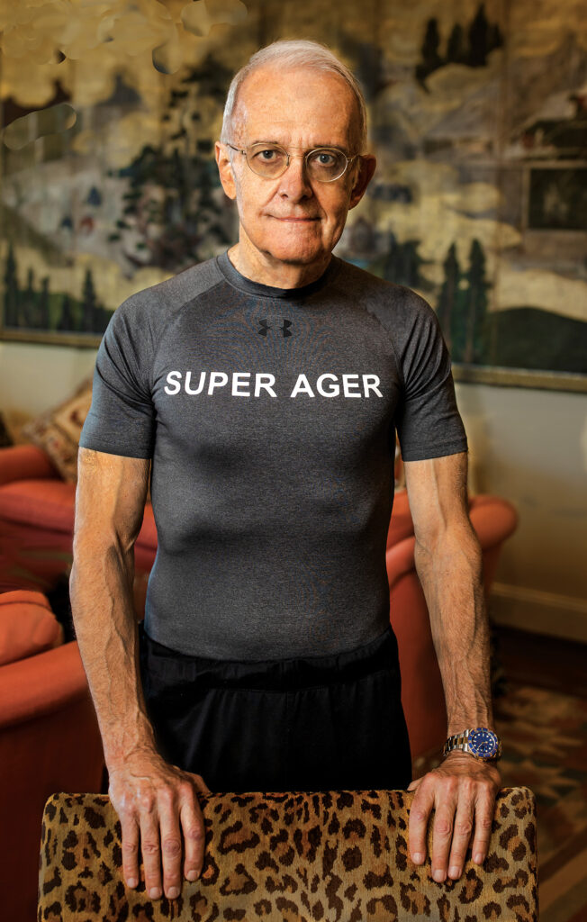 Senior man stands with hands on back of a chair. He wears a gray compression T-shirt with &ldquo;SUPER AGER&rdquo; written across the chest in white
