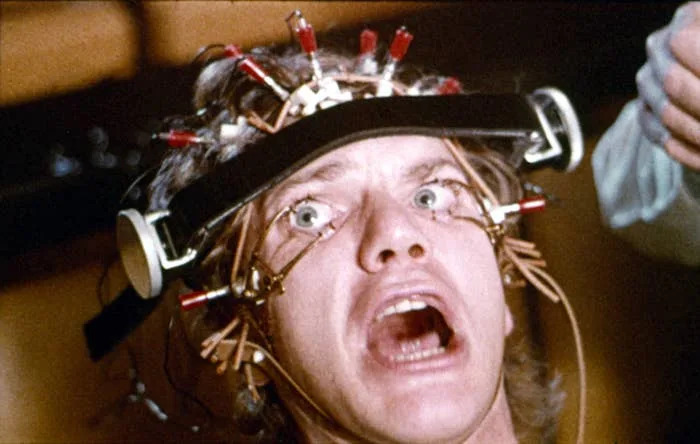 Person with a head apparatus, eyes wide open, undergoing an eye exam, portraying a scene from a movie