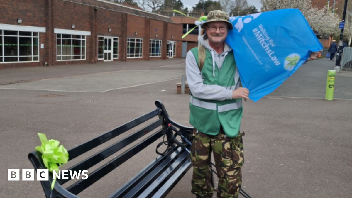 A man wearing a green high-vis jacket, a grey hoodie and camouflage trousers and a hat, holding a blue flag reading "Walking for Mitchs Law" stands next to a black bench with a green bow on it. There is a building behind him