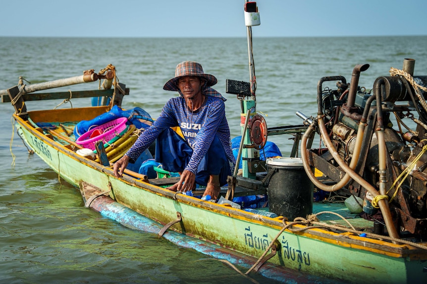 A fisherman squats on the edge of a tiny fishing boat.